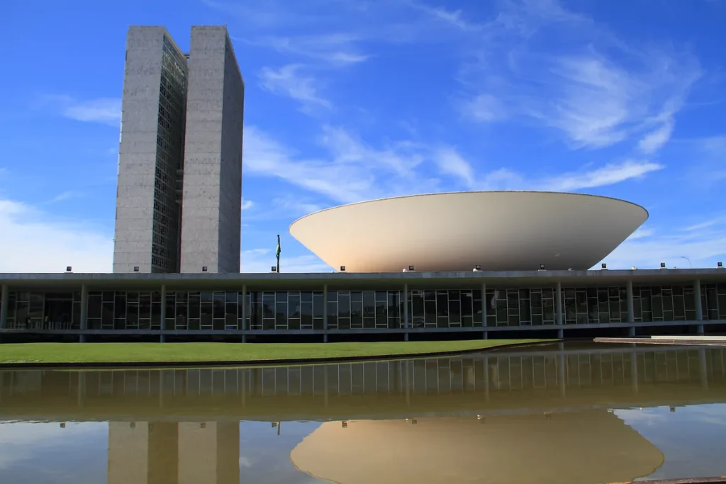 Brazil's National Congress in Brasilia, with its twin office towers beside a white bowl-shaped chamber, reflected in a pool under a blue sky.