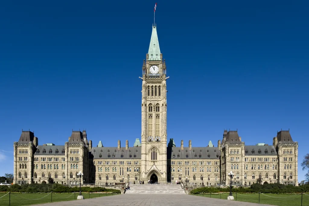 Centre Block on Parliament Hill in Ottawa, the sandstone Gothic Revival building with the Peace Tower and its clock rising from the middle, under a deep blue sky with lawns and a paved walkway in front.
