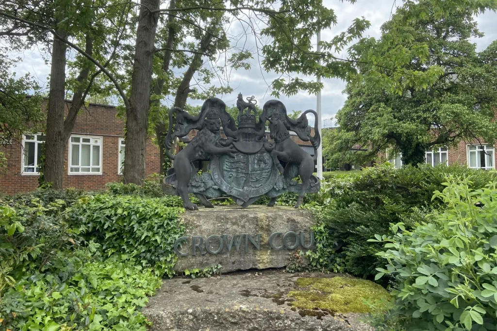 A dark bronze royal coat of arms, a lion and a unicorn either side of a crowned shield, on a stone block carved 'Crown Court', among green shrubs and trees.