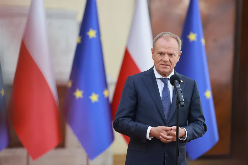 Donald Tusk in a dark suit and blue tie, hands clasped, standing at a microphone in front of Polish and European Union flags.