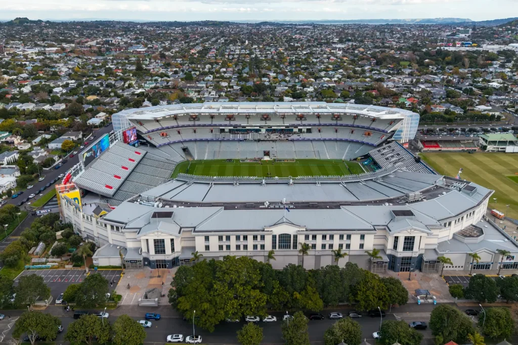 Aerial view of Eden Park stadium in Auckland: white stands around a green rugby pitch, a classical-style grandstand front and tree-lined streets of houses stretching to the horizon.