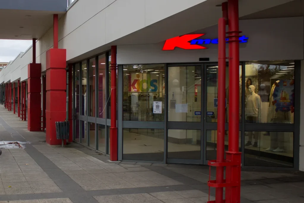 The glass entrance of a Kmart store in a covered shopping arcade, its red and blue sign above the doors and red columns along the walkway.