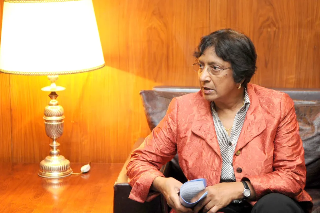 Navi Pillay in a coral jacket and glasses, seated on a dark leather couch beside a brass table lamp against timber panelling, holding a booklet.