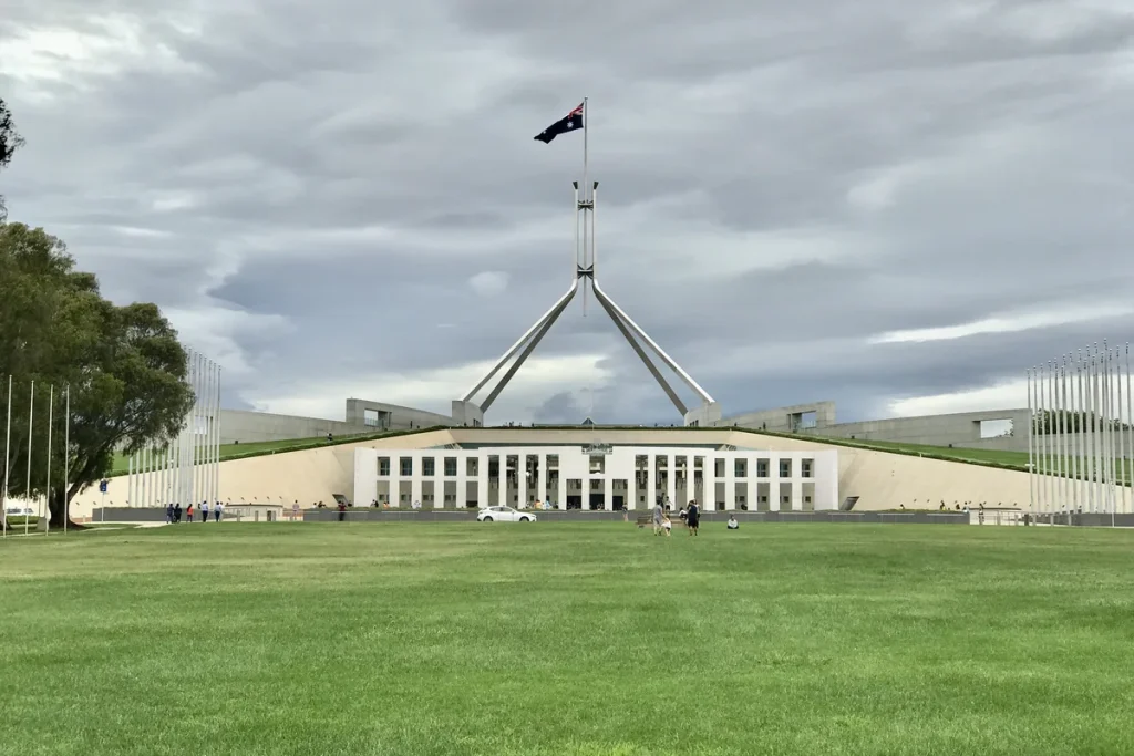 Parliament House, Canberra, under a grey cloudy sky, with the Australian flag flying from the flagpole above the building and a wide green lawn in front. Photographed in December 2016.