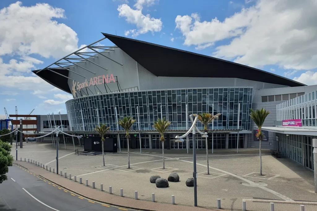 The curved glass and grey-panelled front of Spark Arena in Auckland under a swooping dark roof, with nikau palms and boulders on an empty paved forecourt.