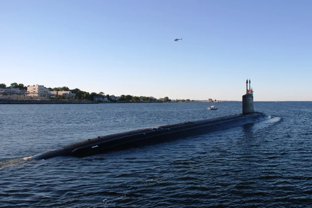 A black Virginia-class submarine running on the surface of the Thames River in Connecticut with its sail and masts raised, a small escort boat, a wooded shoreline with buildings, and a helicopter in a clear sky.