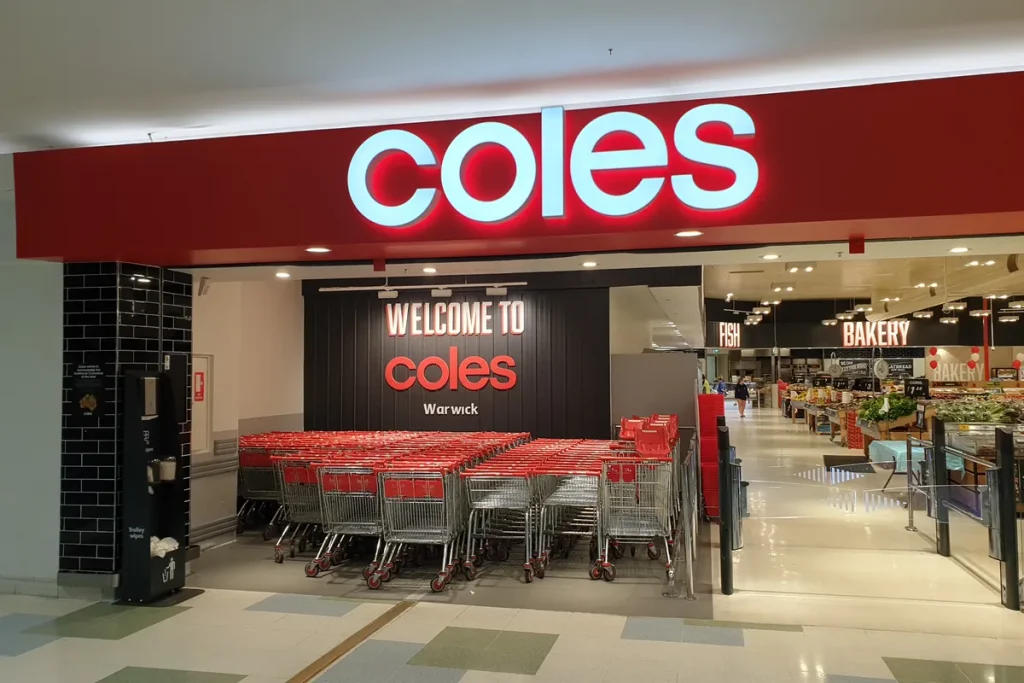 A shopping centre entrance under a red fascia with the Coles name, rows of red-handled trolleys parked inside and fresh produce aisles beyond.