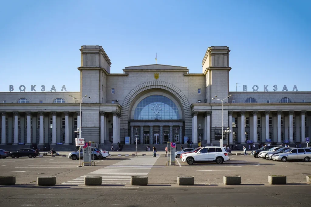 The long pale stone facade of Dnipro's main railway station, with twin towers, a tall arched central window and the word Vokzal in Cyrillic, behind an open car park under a clear sky.