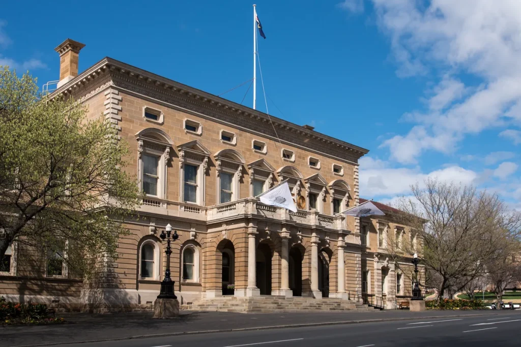 The sandstone Italianate Hobart Town Hall with an arched portico, a balcony with flags and a flagpole on the roof, under a blue sky with spring trees.