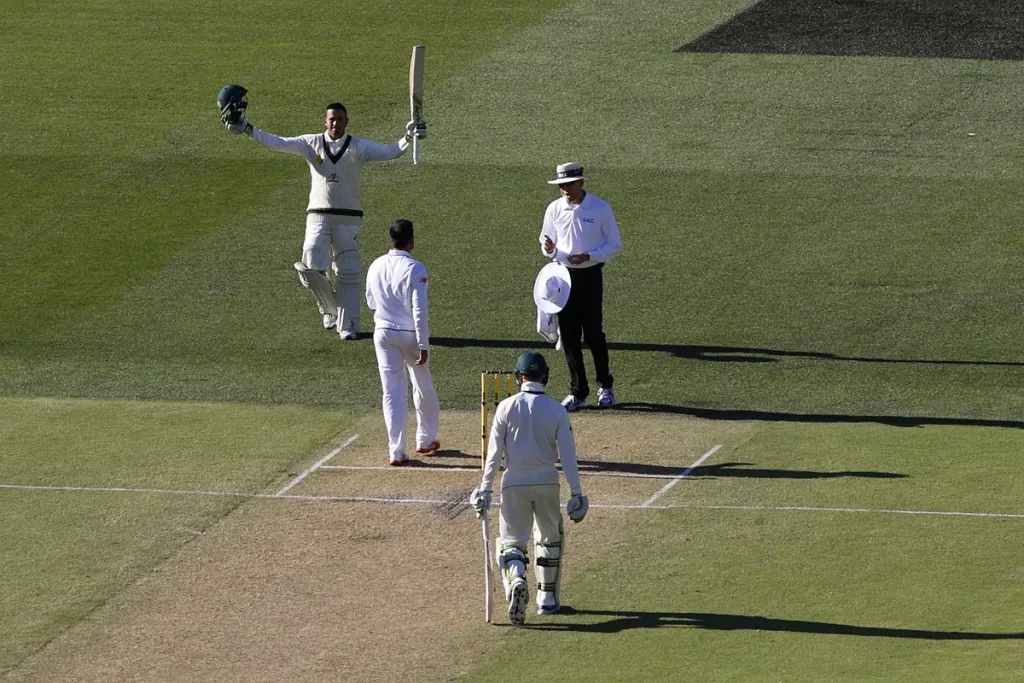 A batter in cricket whites lifts his bat and helmet in celebration on a sunlit Test pitch while an umpire, a fielder and his batting partner stand nearby.
