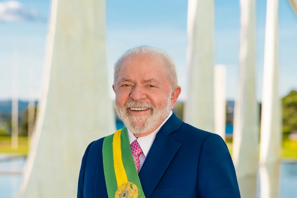 Lula, white-haired and bearded, smiling in a navy suit and pink tie with the green and gold presidential sash, white curved columns behind him.
