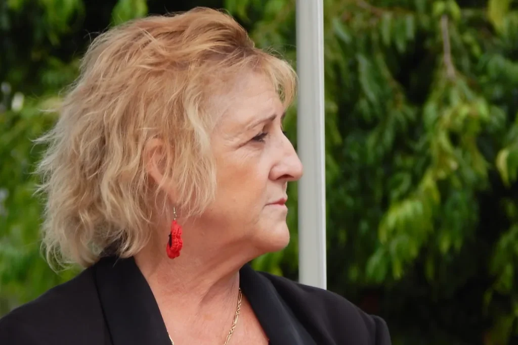 Michelle Landry in profile, wearing a black jacket and red crocheted poppy earrings, with green trees behind her.