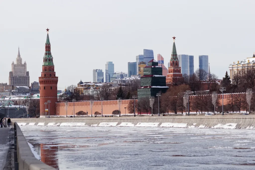 Red Kremlin wall towers topped with stars beside a half-frozen river, with glass skyscrapers rising behind under a pale winter sky.