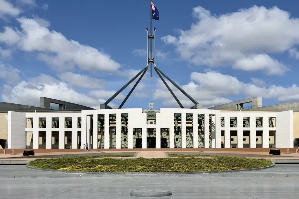 The white colonnaded front of Parliament House in Canberra under a blue sky with clouds, the four-legged flagpole and Australian flag above it.