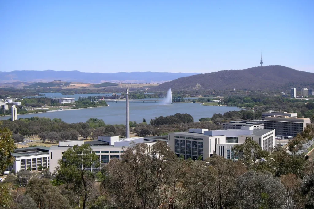 White office blocks of the Russell defence precinct among eucalypts, with the Australian-American Memorial column, Lake Burley Griffin and Black Mountain tower behind.