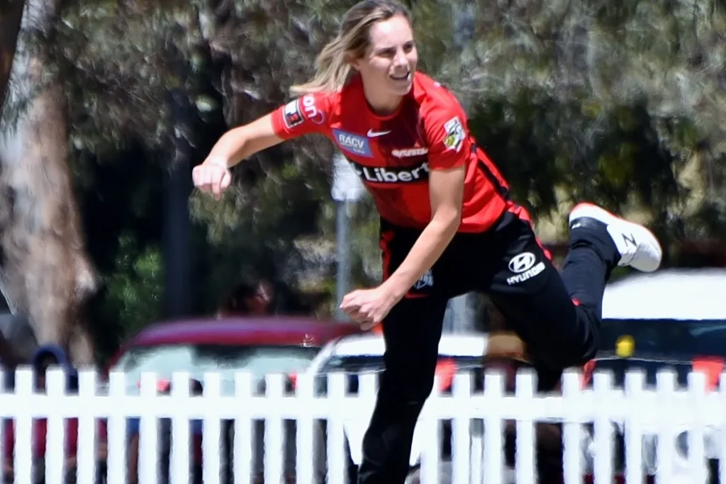 Sophie Molineux in the red and black Renegades kit following through after bowling, a white picket fence and gum trees behind her.