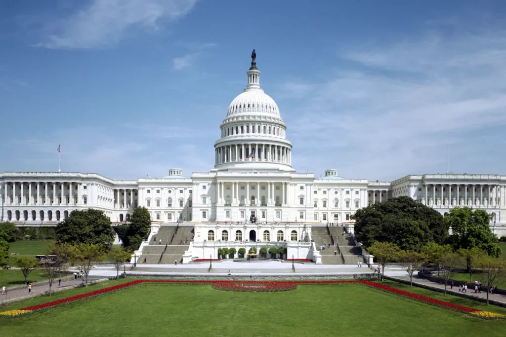 The white domed United States Capitol seen across a green lawn and red flower beds under a blue sky.
