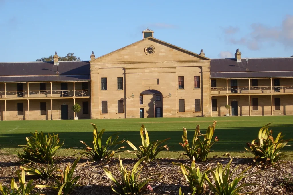 A long two-storey sandstone barrack block with verandahs and a central pediment and clock, behind a parade lawn and a garden bed.
